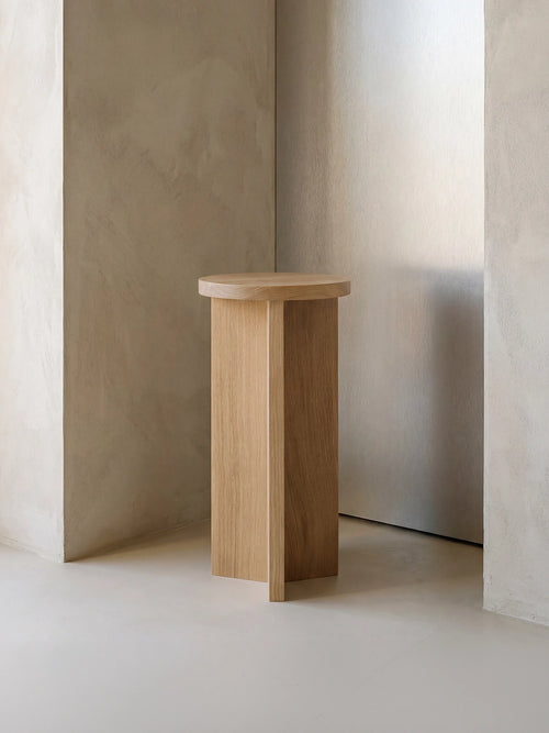 Affe pedestal in solid oak with a round top and sculptural base, styled in a minimalist Scandinavian interior with soft neutral walls and natural light.