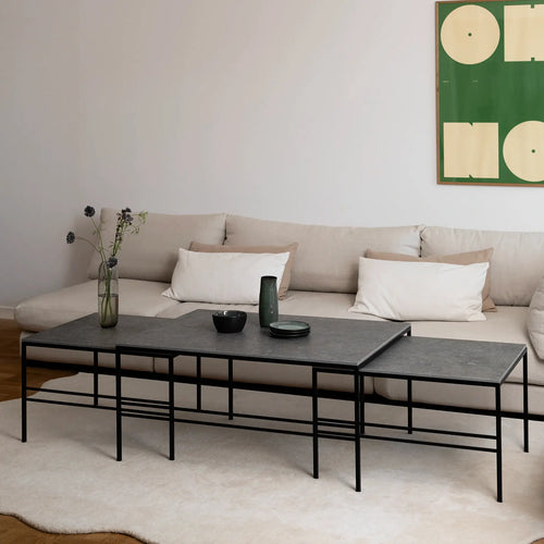 Scandinavian living room showcasing the Anton Coffee Table 3-set by by Crea in Grafite Dekton with a black powder-coated steel frame. The nesting tables are styled with minimalistic green ceramics and a tall glass vase, set on a beige rug in front of a light beige sectional sofa.