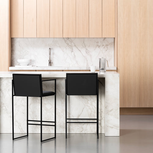 Two modern black By Crea bar stools positioned at a white marble kitchen island, with light wooden cabinetry and a matching marble backsplash in the background. Perfect for interior design, kitchen styling, and furniture e-commerce.