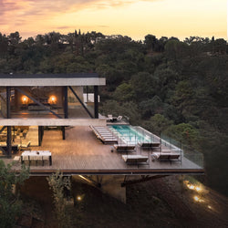 Architectural hillside villa with a spacious wooden deck at sunset, showcasing a line of Bazo Sunbed Loungers surrounding an infinity pool, overlooking a forested valley.