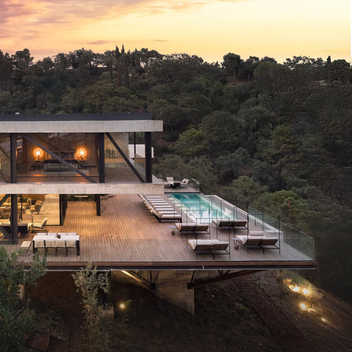 Architectural hillside villa with a spacious wooden deck at sunset, showcasing a line of Bazo Sunbed Loungers surrounding an infinity pool, overlooking a forested valley.
