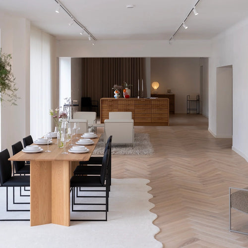 Minimalist Scandinavian showroom by Crea featuring a natural oak dining table set with black-framed chairs, cream lounge seating, and curated decor, all in a soft-toned herringbone space.
