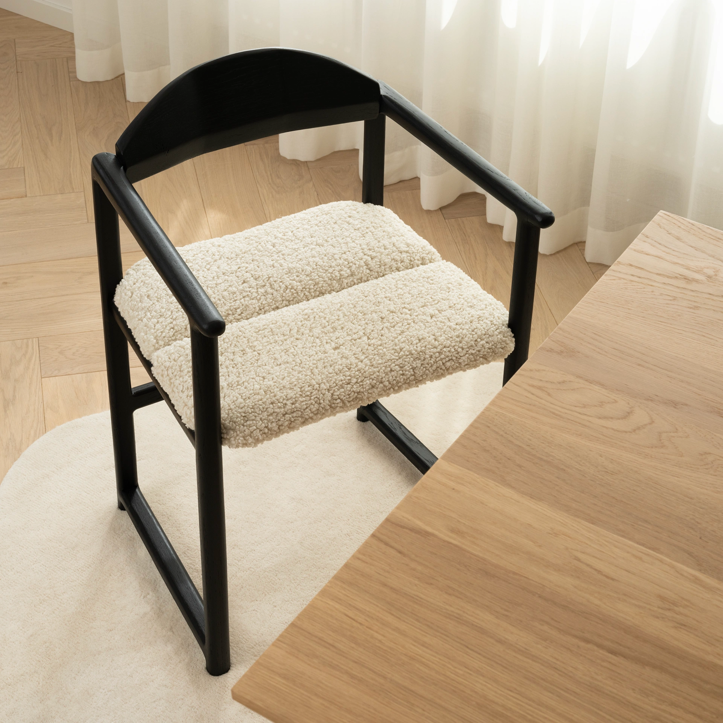 Close-up top view of the Erik dining chair in black oak with ivory bouclé cushion, paired with a solid oak table on a light rug in a Scandinavian interior.