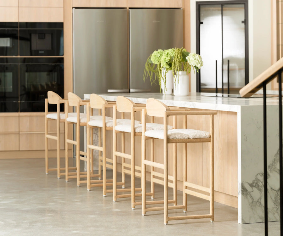 Carl Bar Stools in natural oak with ivory bouclé cushions, neatly aligned along a waterfall-edge marble kitchen island in a Scandinavian-style home with light wood cabinetry and polished concrete floors.