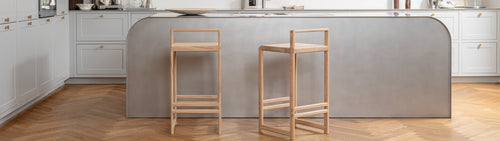 Ebba Bar Stools in bright oak placed at a minimalist grey kitchen island with curved edges, set against white shaker-style cabinets and herringbone wooden flooring.