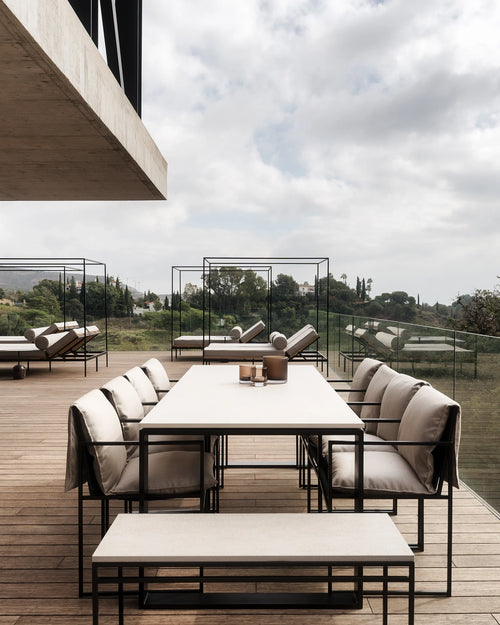 Minimalist outdoor dining area with the Diana Outdoor Table in Caesarstone, surrounded by black-framed armchairs with soft beige cushions, and matching bench seating on a wooden deck with panoramic views.