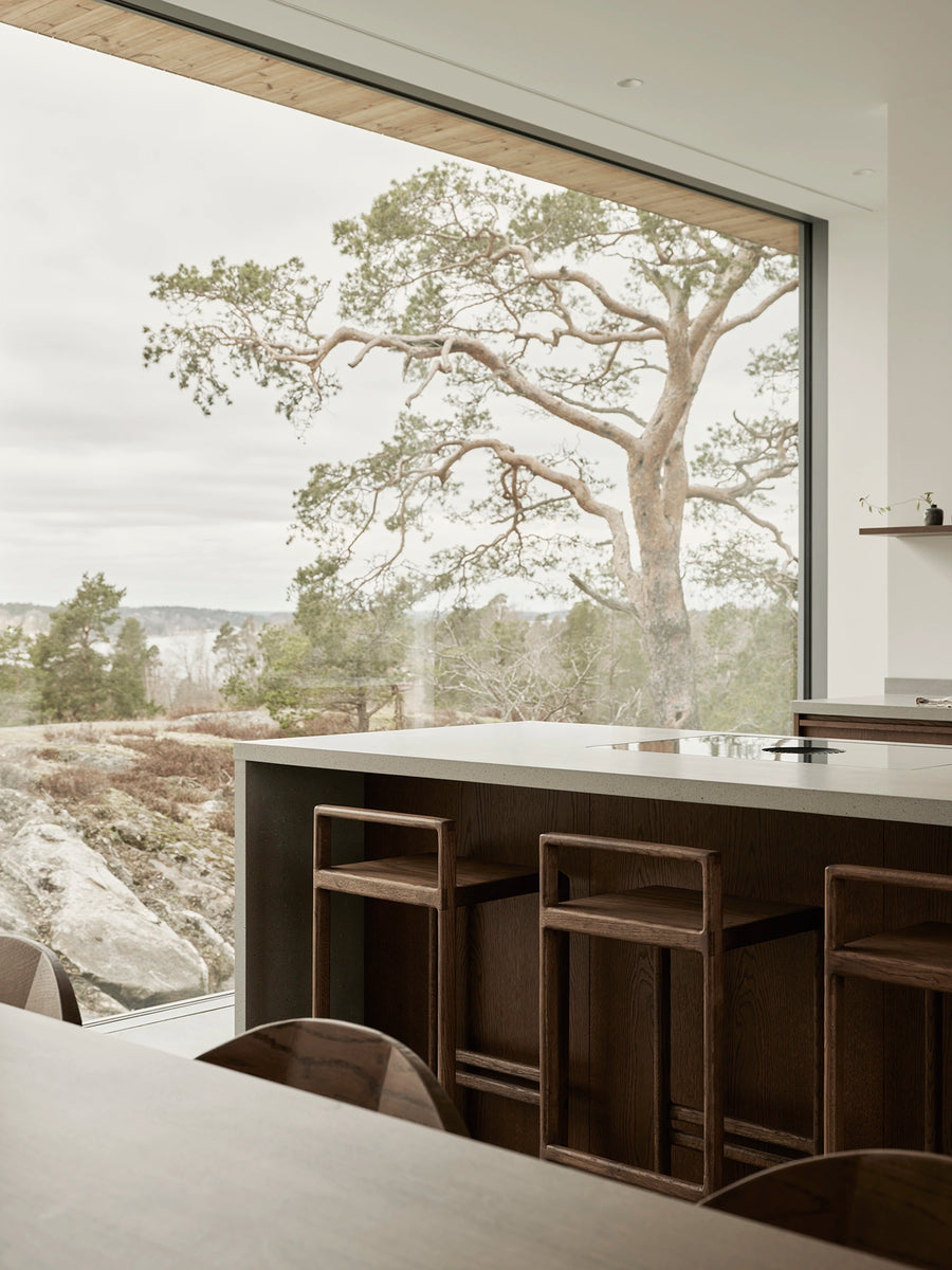 Minimalist Scandinavian kitchen with oak bar stools at an island, framed by natural light and a landscape view.