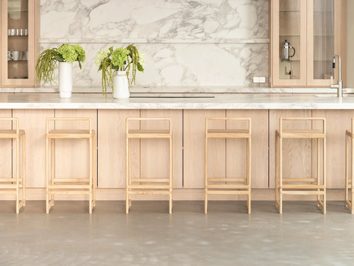 Ebba Bar Stools in natural oak lined up at a Scandinavian-style kitchen island, featuring a marble countertop and backsplash with light wood cabinetry and minimalist floral decor.