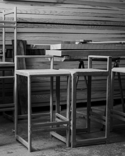 Unfinished Ebba bar stools in raw solid oak at the by Crea factory, surrounded by stacked timber and workbenches. Captures the craftsmanship of Scandinavian handmade furniture in progress.