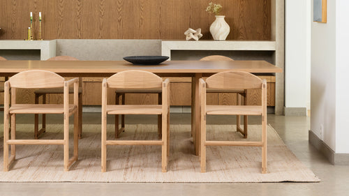 Minimalist dining room featuring six Erik dining chairs in natural oak with clean lines and joinery details, paired with a matching wood table and neutral rug, set against a wood-paneled and concrete niche backdrop.