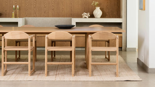 Minimalist dining room featuring six Erik dining chairs in natural oak with clean lines and joinery details, paired with a matching wood table and neutral rug, set against a wood-paneled and concrete niche backdrop.