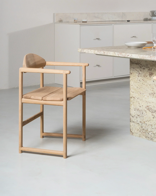 Erik Dining Chair by by Crea in solid oak with armrests, placed beside a stone countertop in a minimalist kitchen interior.