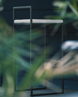 Minimalist Freja outdoor bar stool with sleek black frame and cushioned white acrylic seat, set against a dark exterior wall with soft greenery in the foreground.