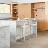 Freja Bar Stools in beige with minimalist frames, positioned at a grey-veined marble kitchen island in a light oak kitchen with open shelving.