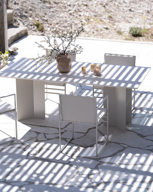 Sunny outdoor dining area featuring a sleek by Crea Ann rectangular outdoor table and matching chairs in a light beige or off-white finish, with sturdy block legs, placed on an irregularly paved stone patio, with strong striped shadows from an overhead pergola or trellis, and a vase of white blossoms as a centerpiece.