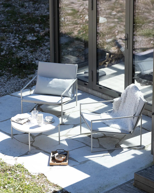 Outdoor patio setup featuring two by Crea Kent lounge chairs with sleek light grey frames and matching cushions, one draped with a white faux fur throw, beside a minimalist by Crea Leo lounge table holding a glass teapot, mugs, and a tray, set on light grey flagstones next to glass doors.