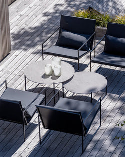 Outdoor patio on a sunny day featuring four by Crea Kent lounge chairs with sleek black metal frames and dark grey cushions, arranged around a modular by Crea Leo lounge table set consisting of two round grey tabletops, resting on a weathered wooden plank deck.