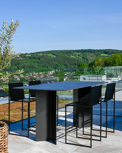 Sleek outdoor dining area on a high-end rooftop terrace, featuring four by Crea Philip bar stools with black frames and minimalist design, paired with a large rectangular black outdoor bar table, set on light grey paving with green roof elements and a glass railing, offering panoramic views of rolling green hills and a town below