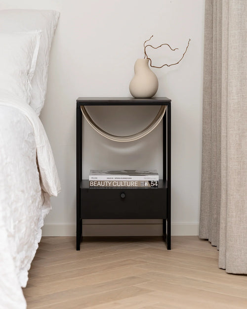 Stylish Iris nightstand with black metal legs, a lower pull-out drawer, and unique arched fabric sling for books, positioned beside a white bed with light wood flooring.