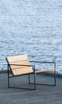 Kenny Outdoor Lounge Chair by by Crea, featuring teak wood seat and black steel frame. Pictured alone on a wooden dock with calm blue water in the background, highlighting minimalist Scandinavian design and tranquil outdoor living.