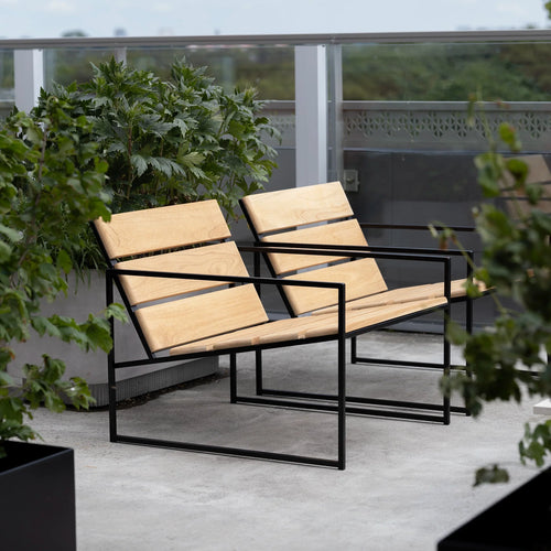Kenny Outdoor Lounge Chair in teak wood with black steel frame by by Crea, minimalist Scandinavian design. Shown on a modern balcony with lush green plants and concrete flooring, ideal for urban outdoor living, handcrafted in Sweden.