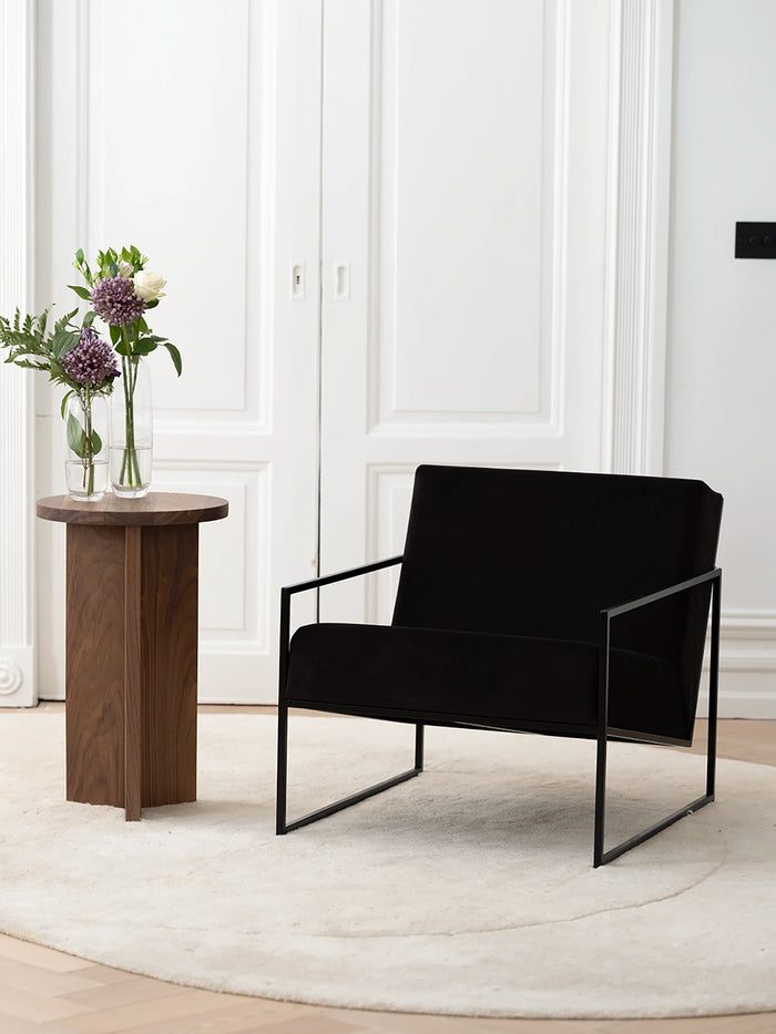Armchair Kent in black velvet with a sleek black frame, paired with a wooden side table in a stylish minimalist setting.