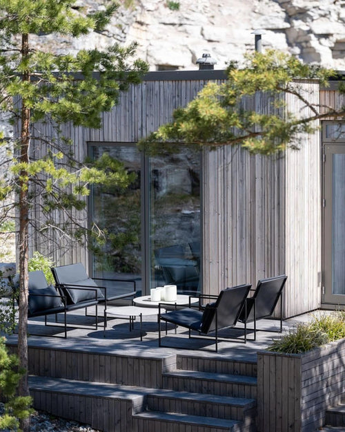 Set of black Kenny outdoor lounge chairs with sleek steel frames and low-profile design, styled on a modern Scandinavian wooden deck with a round table and minimalist candle decor, surrounded by nature.
