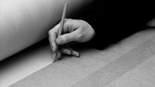 Black-and-white image of a craftsman’s hand tracing upholstery fabric using a pencil and paper stencil, highlighting the artisanal process behind by Crea’s handmade Scandinavian furniture.