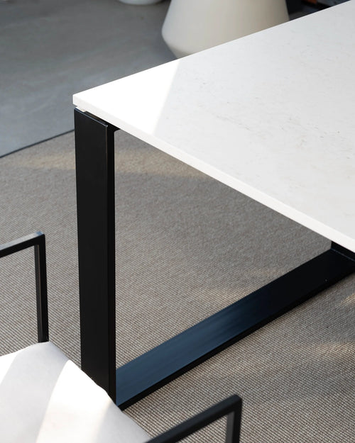 Detailed view of the sleek by Crea Diana outdoor dining table, highlighting the crisp white tabletop, possibly made of a durable composite or stone, contrasted with its strong, minimalist black powder-coated steel U-shaped base, set on a textured grey outdoor rug.