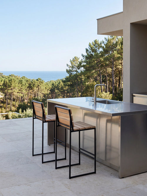 Modern outdoor kitchen with stainless steel island and stools, surrounded by trees and open sky.