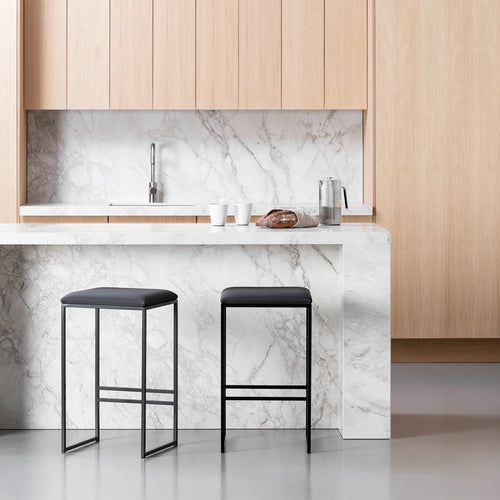 Two By Crea Svea all-black bar stools with sleek metal frames and dark seats, positioned at a white marble kitchen island, with light wooden cabinetry and a marble backsplash in the background. Ideal for modern minimalist kitchen designs and contemporary furniture.
