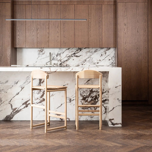 Minimalistic solid oak Ted bar stools in a modern kitchen with marble countertop and wooden cabinets.