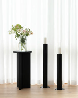 Thomas Candle Holder 2-set in black steel with glass covers styled alongside the Affe Pedestal in black oak and a ribbed glass vase with fresh flowers, set against sheer white curtains in a minimal Scandinavian interior.