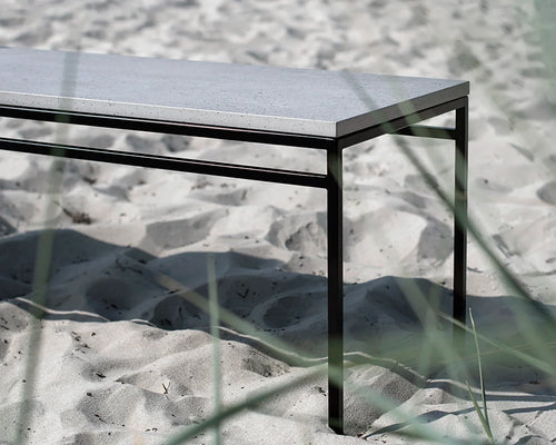 Wilma outdoor bench with a black powder-coated steel frame and durable Dekton stone top, styled in a coastal sand setting with natural grass in the foreground.