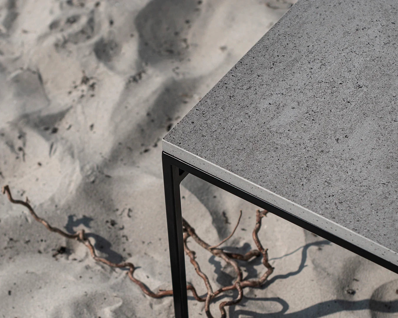 Detail view of the Wilma outdoor bench showing its black powder-coated steel frame and grey Dekton stone top, set against a sandy beach backdrop.