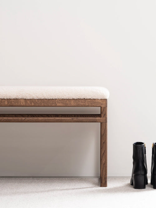 Oak Bench Alva with dark oak frame and ivory bouclé cushion, beside stylish black boots.