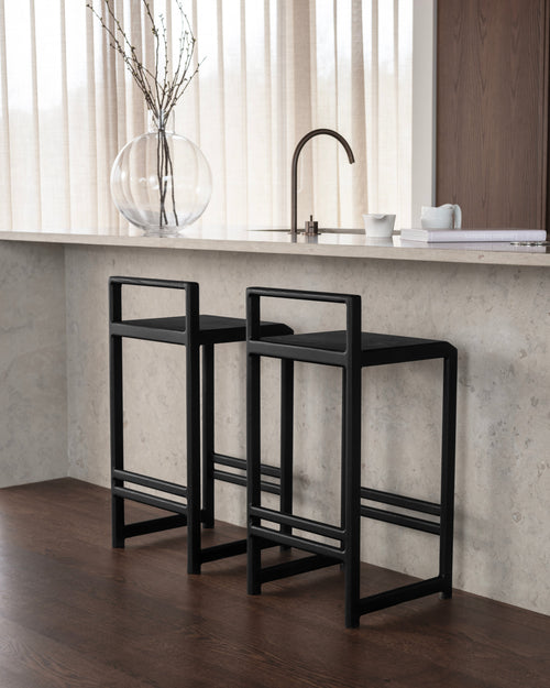 Ebba Bar Stools in black oak by Crea styled at a stone kitchen island with dark wood flooring and neutral curtains.