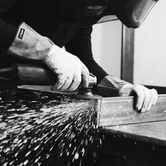 Close-up black and white image of a craftsman in protective gear grinding a metal frame, with visible sparks flying, highlighting the handmade production process at by Crea