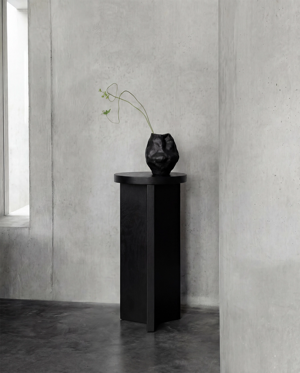 Affe pedestal in black-stained solid oak with a round top and sculptural base, placed against raw concrete walls in a minimalist Scandinavian interior with a black ceramic vase on top.