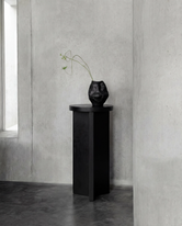 Affe pedestal in black-stained solid oak with a round top and sculptural base, placed against raw concrete walls in a minimalist Scandinavian interior with a black ceramic vase on top.