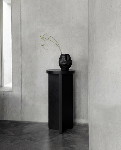 Affe pedestal in black-stained solid oak with a round top and sculptural base, placed against raw concrete walls in a minimalist Scandinavian interior with a black ceramic vase on top.