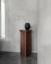Affe pedestal in dark oak with visible natural wood grain, styled against raw concrete walls in a minimalist Scandinavian interior with a sculptural black vase on top.