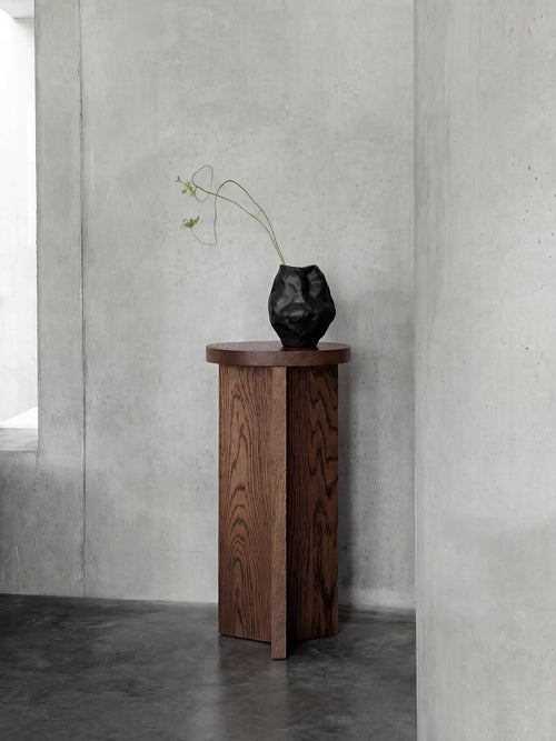 Affe pedestal in dark oak with visible natural wood grain, styled against raw concrete walls in a minimalist Scandinavian interior with a sculptural black vase on top.
