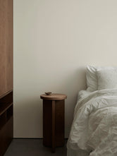 Solid oak pedestal side table beside a bed in a calm, minimalist Scandinavian bedroom interior.