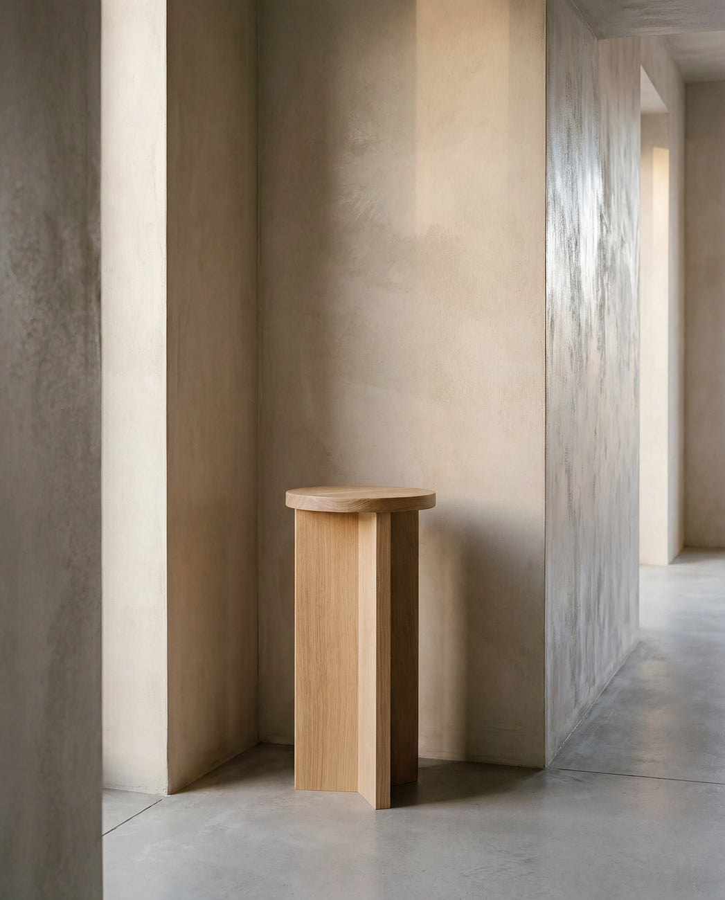 Affe pedestal in solid oak with a round top and architectural base, placed in a minimalist Scandinavian interior with textured neutral walls and soft natural light.