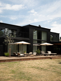 Black timber villa in Höllviken with by Crea outdoor furniture along the deck: Arvid lounge chairs, Bazo sun loungers and cream parasols against the dark facade.
