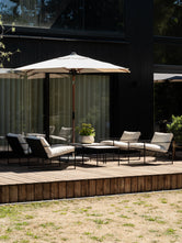 Modern outdoor lounge setting on a raised timber deck, featuring black metal-framed Kenny lounge chairs with cream cushions, coordinating low tables and a large cream parasol against the striking façade of a black-clad contemporary home.