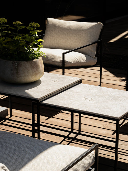 Close-up of nested Anton outdoor lounge tables with light Sabbia stone tops and black metal frames, paired with Kenny lounge chairs in textured light oat cushions on a sunlit timber deck.