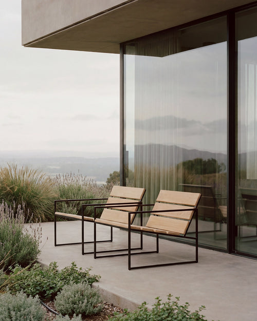 Outdoor lounge chairs with black steel frames and wooden slats placed on a minimalist terrace, styled against a modern glass façade with soft landscaping and scenic natural views.