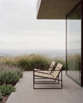 Side view of an outdoor lounge chair with a black steel frame and wooden slats, placed on a minimalist terrace with soft landscaping and expansive natural views beside a modern glass facade.